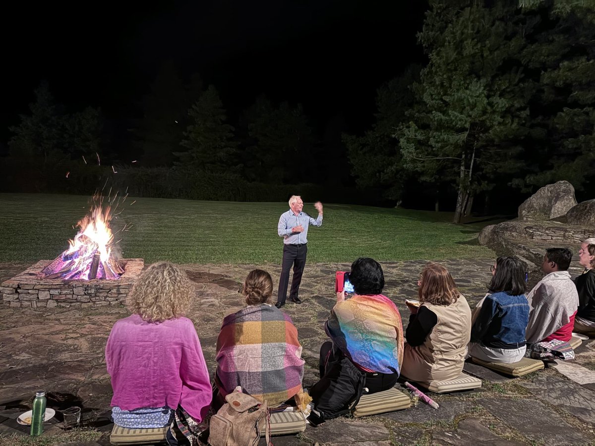 Participants seated on cushions around an open fire at night, listening to a speaker