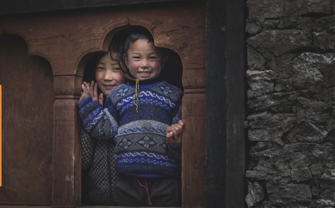 Two smiling Bhutanese children leaning out of a carved wooden window