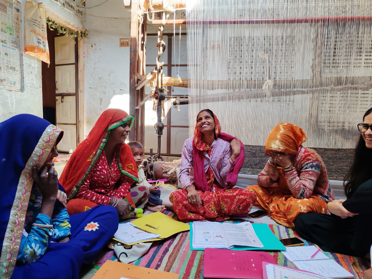 Women artisans sit in a circle on a striped rug beside a carpet loom in rural Rajasthan, laughing together with workbooks open between them.