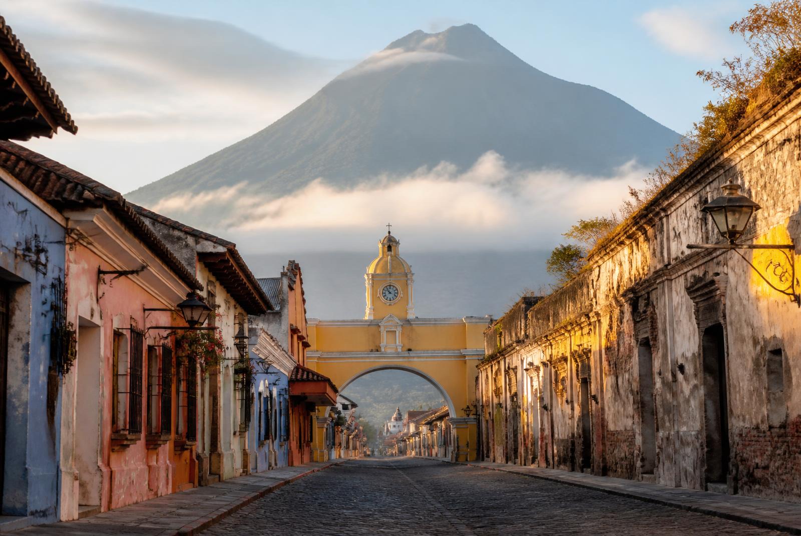 Calle empedrada de Antigua Guatemala con el Arco de Santa Catalina y un volcán al fondo.