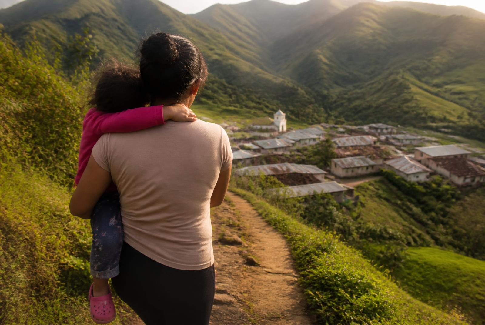 A caregiver carrying a child on a mountain path above a village at golden hour